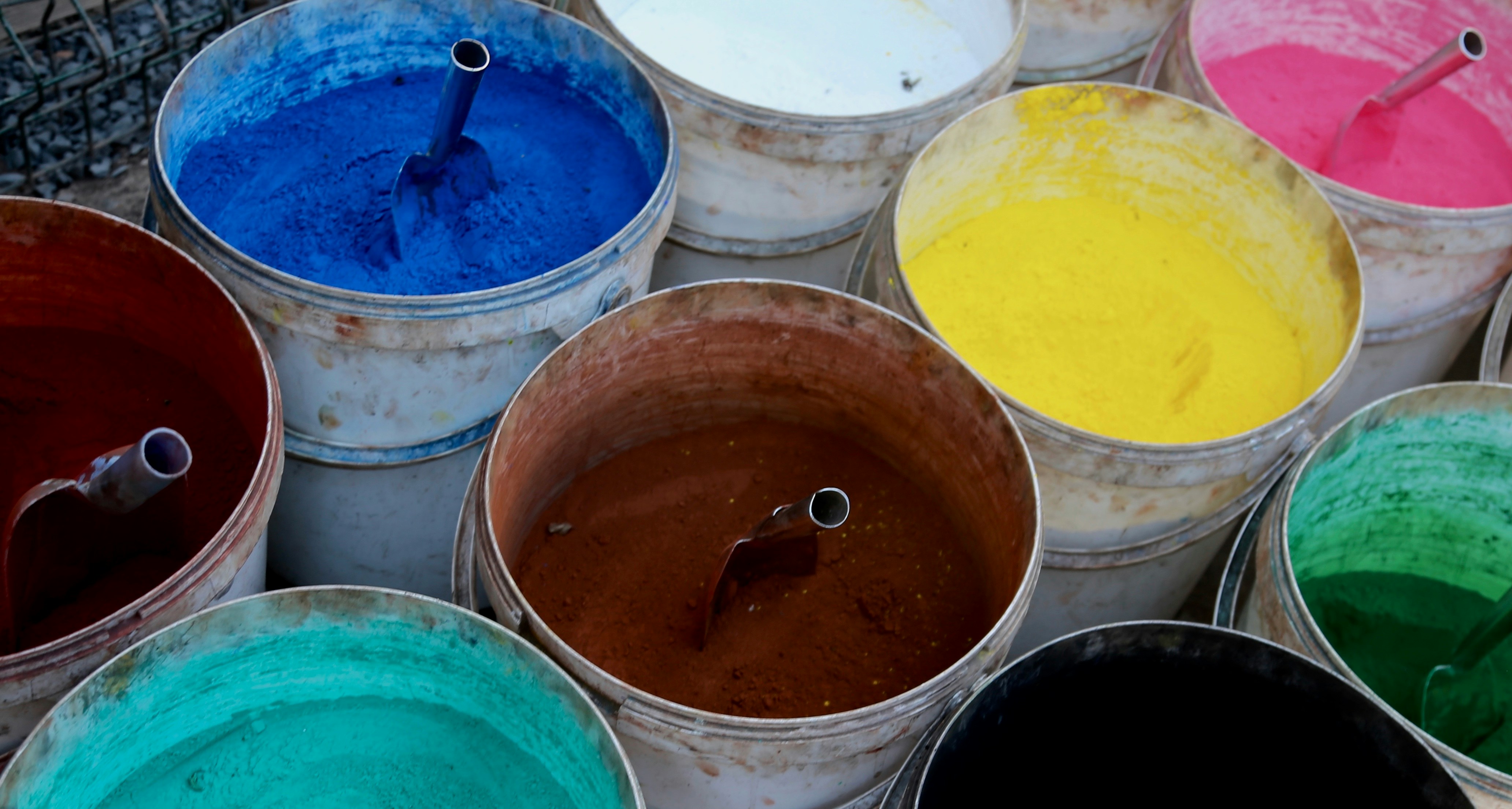 A photo of various colored powdered dyes in white bucket. Silver metal scoops are in each bucket.