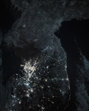 A black and white picture of the Korean Peninsual. Areas of bright light with webs of light connecting them to smaller areas cover the South, while in the north only two small areas of light can be seen.