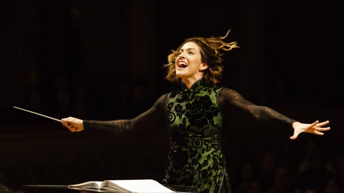 Conductor Alondra de la Parra wearing a green and black chinese-style tunic, her arms outstretched as she conducts with a live audience in the background