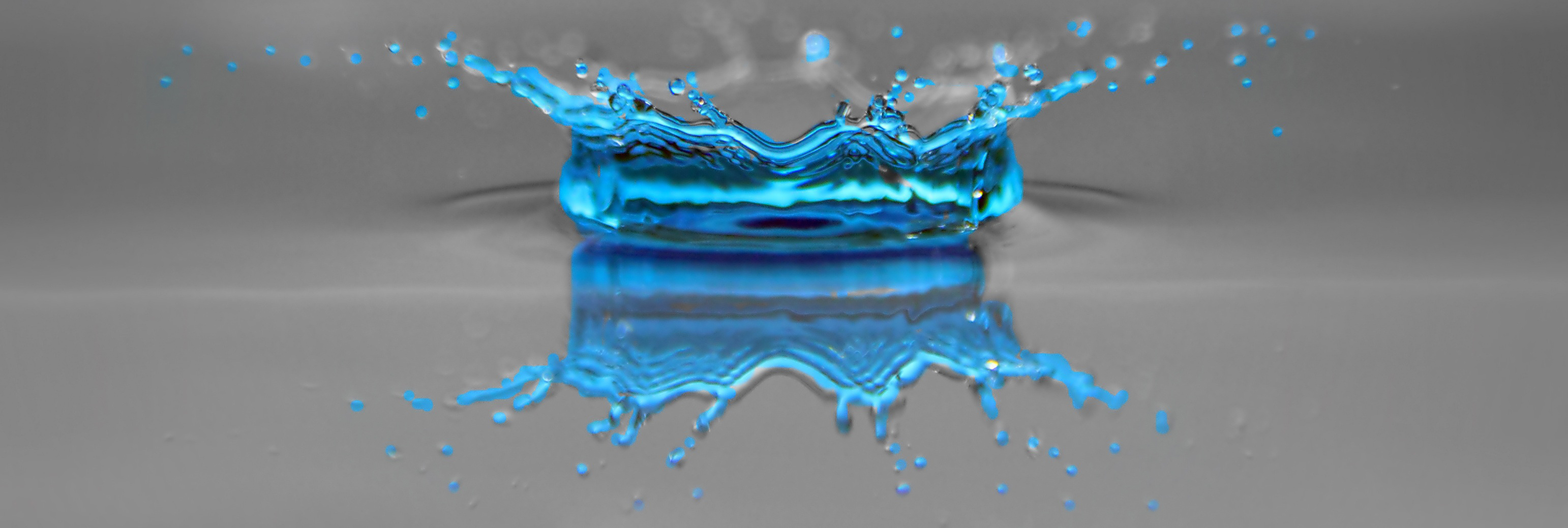 An intense closeup of a water droplet in blue, splashing in a reflected crown shape, against a grey background