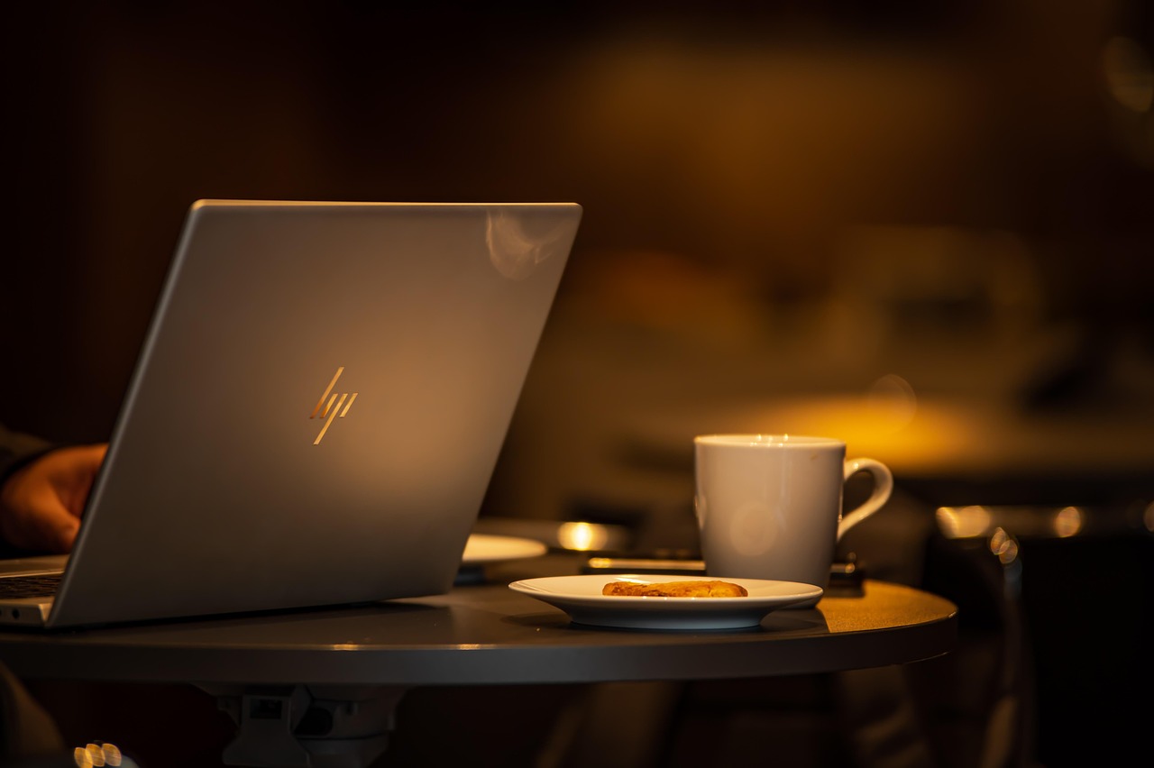 laptop-7956356_1280.jpg A laptop on a table in a dimly lit coffee shop, with coffee in a mug and food on a plate