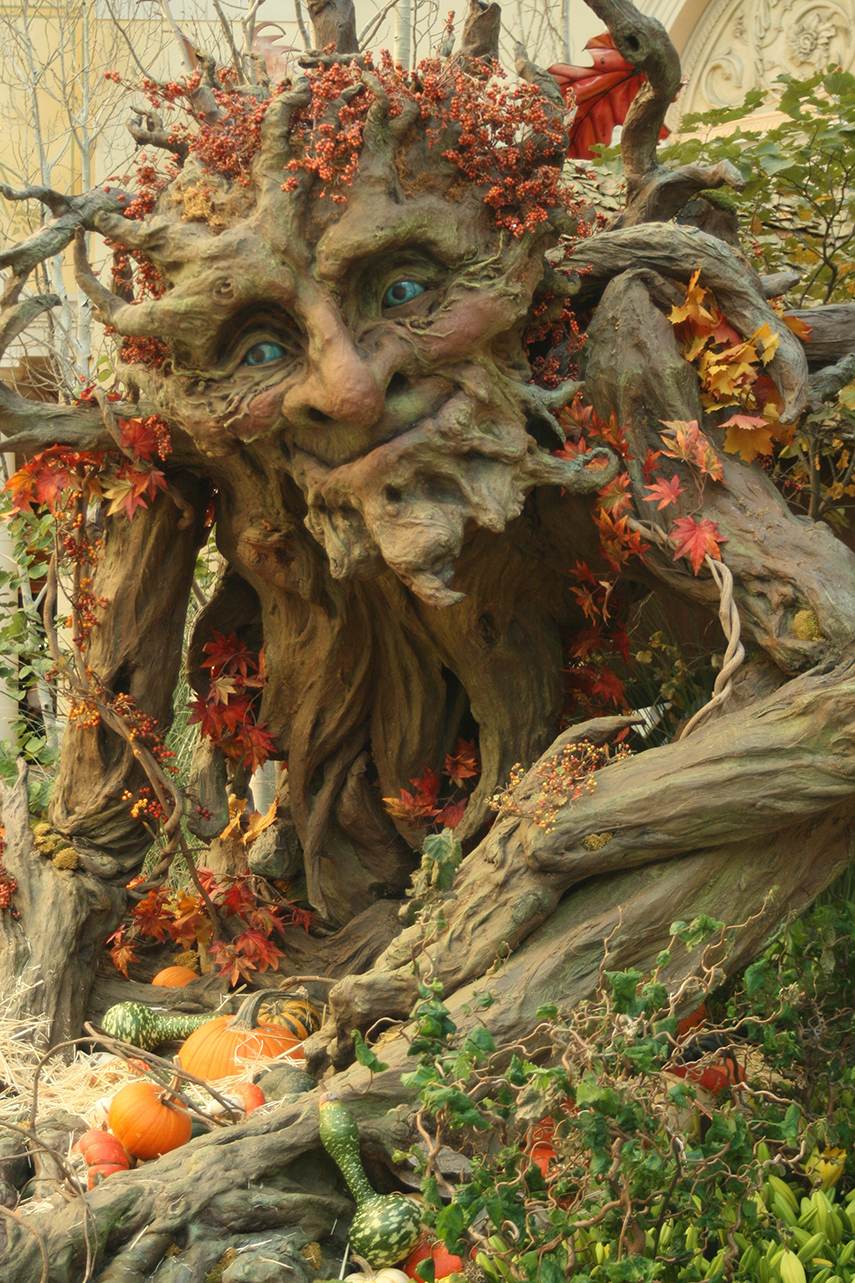 A large, smiling tree man with blue eyes, decorated with autumn leaves and gourds