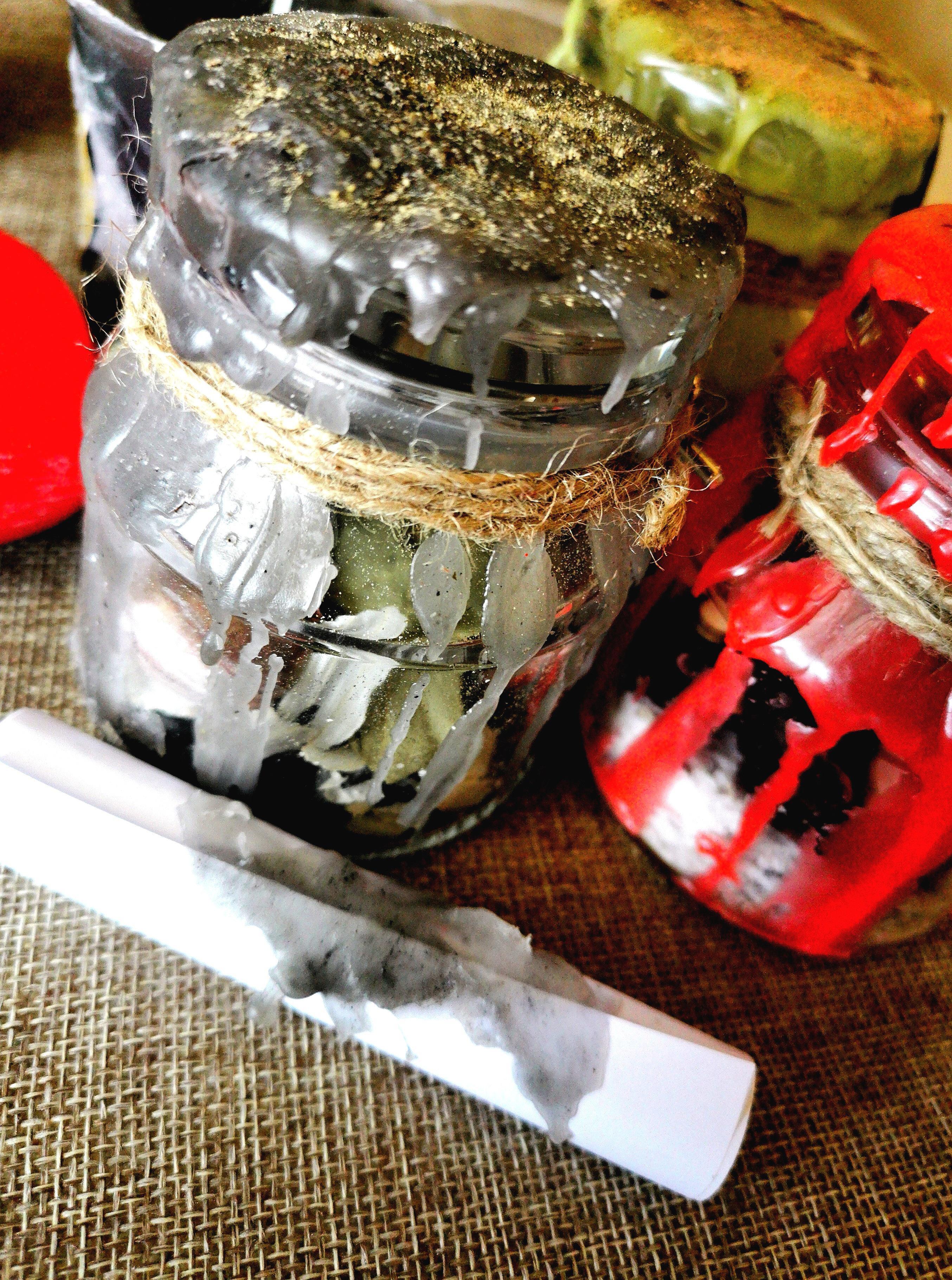 Two wax and glitter-covered canning jars, filled with herbs & other ingredients, on burlap, with a wax-stained rolled paper in front