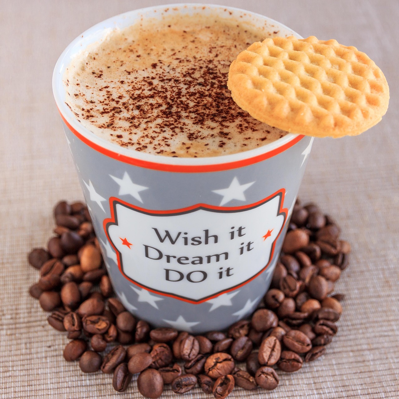 coffee-1587079_1280.jpg A coffee cup, surrounded by coffee beans, filled with a foamy chocolate-sprinkled beverage, with a waffle on the rim of the cup. Cup design is grey with red highlights, and features star designs and text: